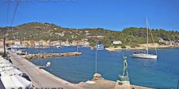 Webcam Gaois - Paxos Harbor with sailboats on blue water beside a stone pier and hillside village under a clear sky
