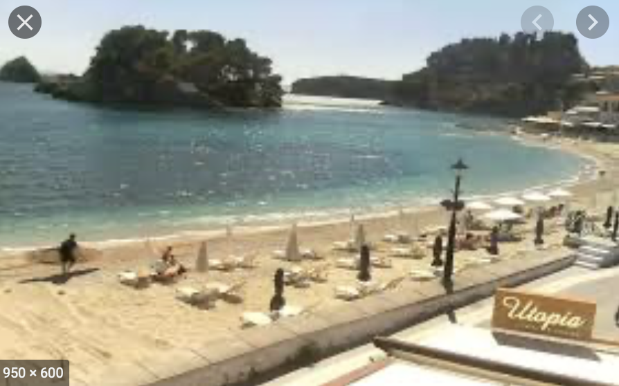 Webcam Parga Beach with turquoise water, lounge chairs, umbrellas, and a small island in the background