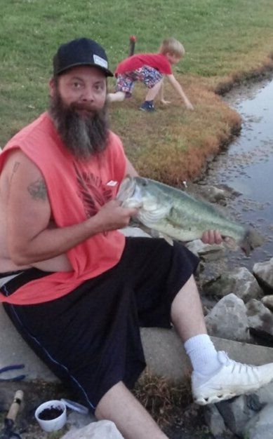 Man holding a large bass fish; boy in background on grassy bank.