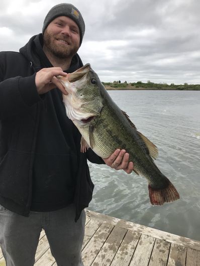 Man holding a large bass fish on a dock, smiling. Cloudy day over water and land.