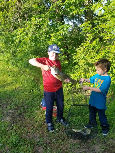 Two boys hold up a large fish, standing in front of green foliage and grass.