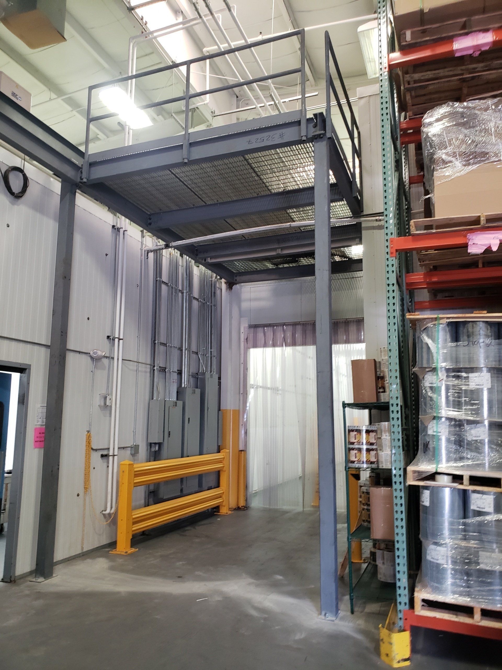 Warehouse with a gray mezzanine platform, safety railing, and yellow barrier. Shelving and a loading bay are visible.