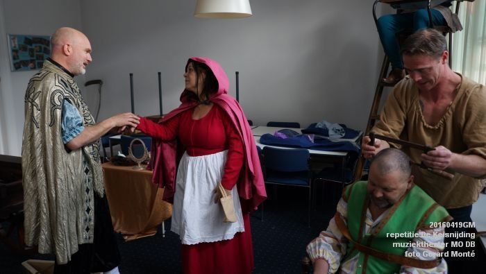 Acteurs in kostuum op een set. Een vrouw in een rode mantel praat met een man in een gouden cape, terwijl anderen zich voorbereiden op de opnames.