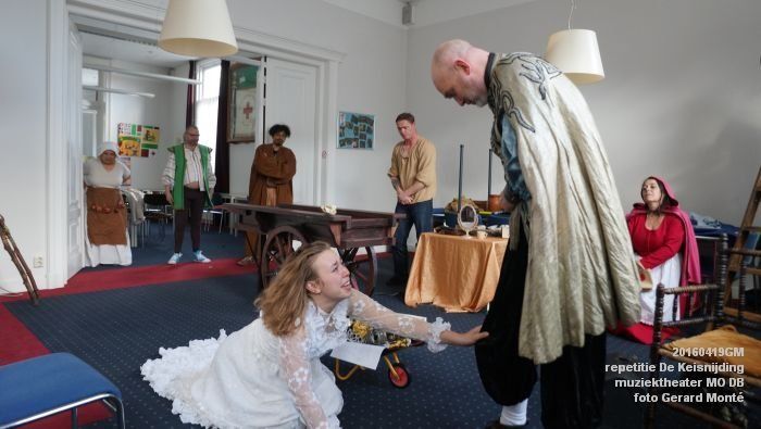 Een theatrale scène met acteurs in kostuum. Een vrouw knielt en reikt naar het been van een man. Andere acteurs kijken toe.