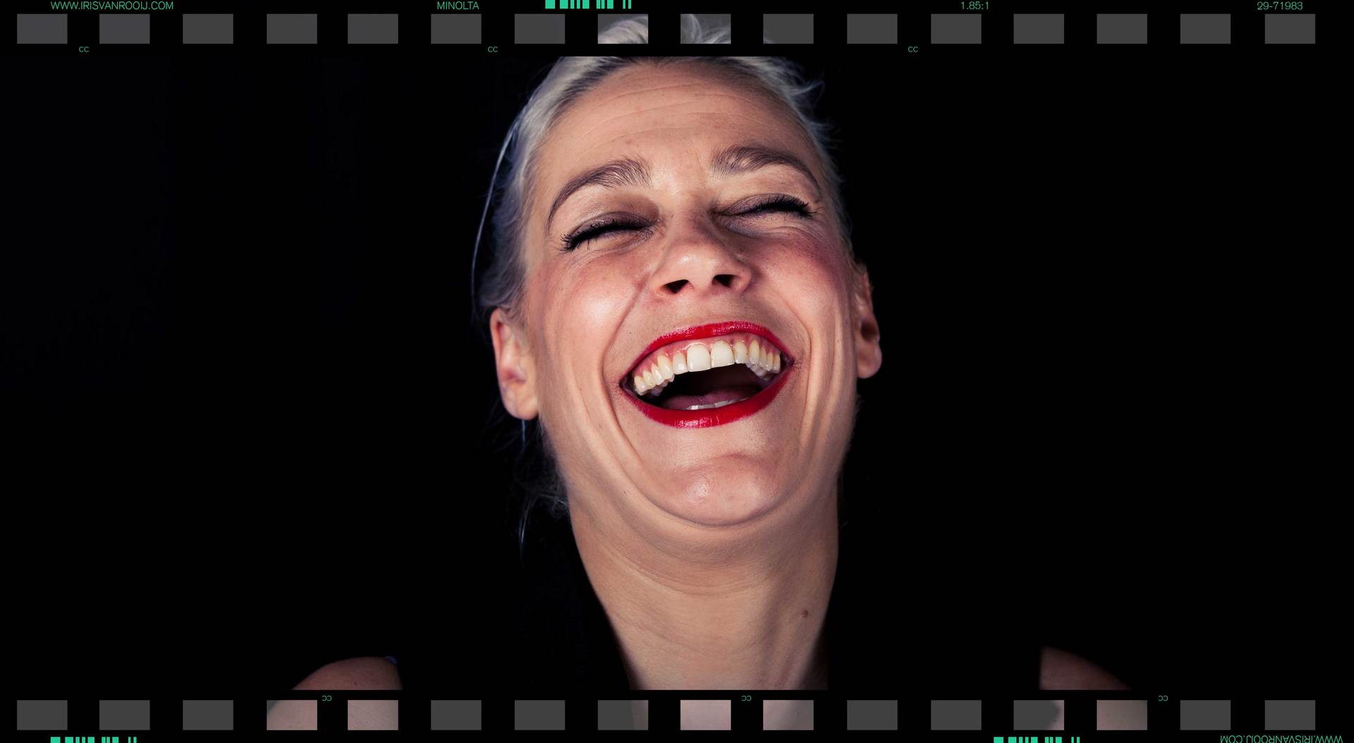 Vrouw met zilverkleurig haar lacht, ogen gesloten, rode lippenstift tegen een zwarte achtergrond.