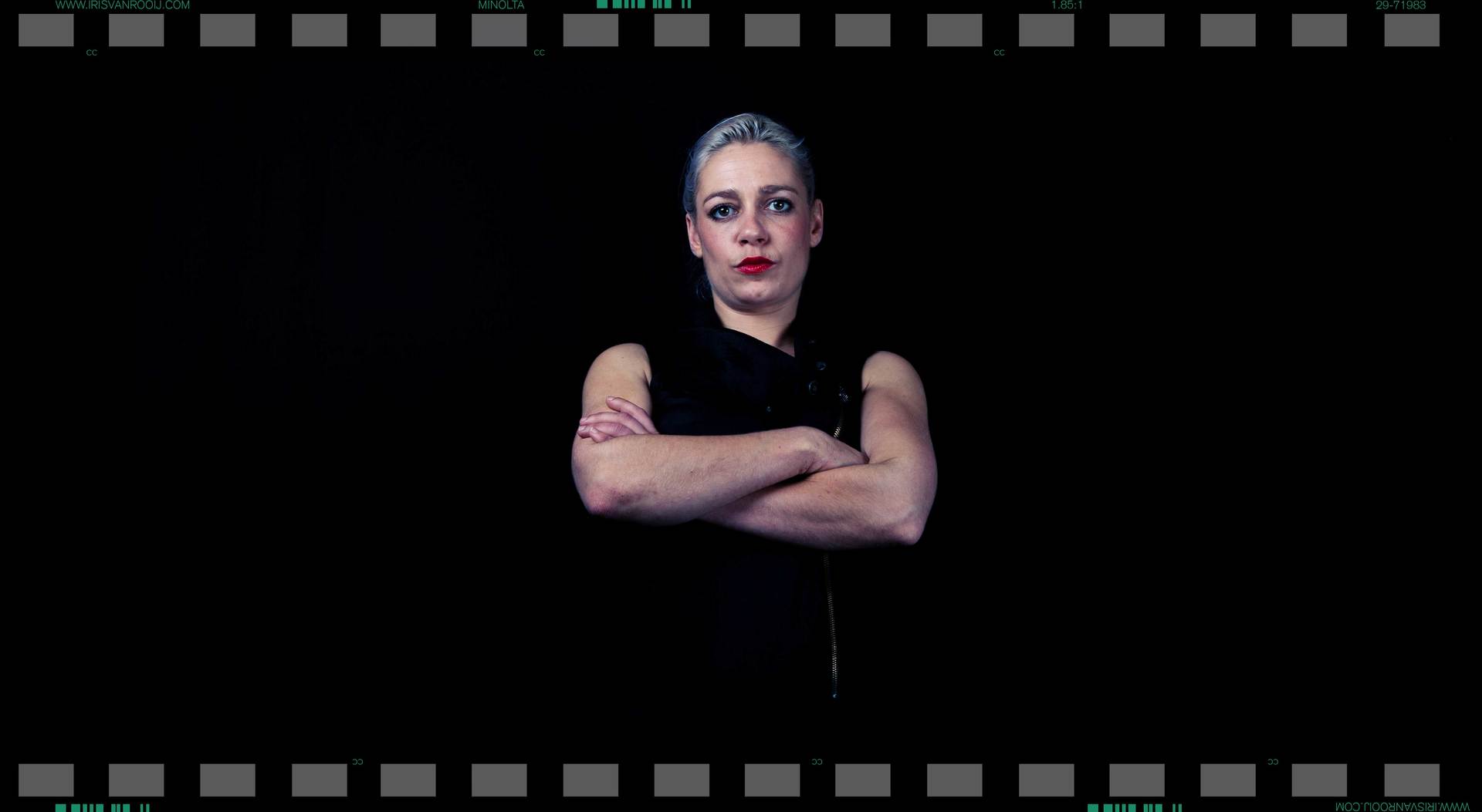Vrouw met kort, zilvergrijs haar, armen over elkaar, gekleed in een zwarte top, tegen een zwarte achtergrond.