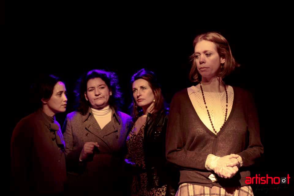 Vier vrouwen op het podium; de vrouw op de voorgrond, gekleed in een donkere trui en geruite broek, staat in de spotlight. Achter haar staan ​​drie vrouwen in verschillende kleding.