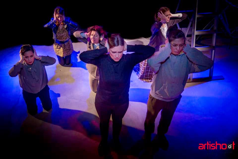 Mensen op het podium houden hun oren dicht. Gedempte verlichting, theatrale setting.