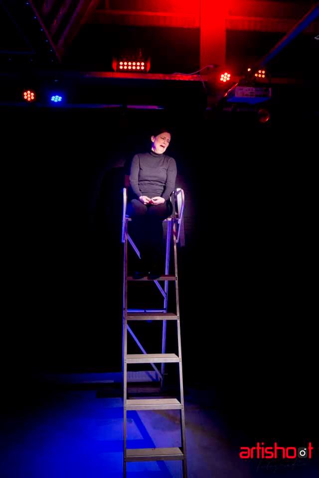Een persoon zit op een ladder onder podiumverlichting. Donkere omgeving.