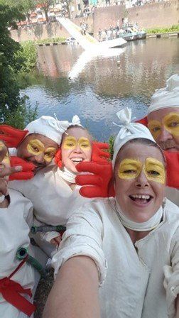 Mensen in witte kostuums, met gele oogmake-up en rode handschoenen, poseren voor een selfie bij het water.