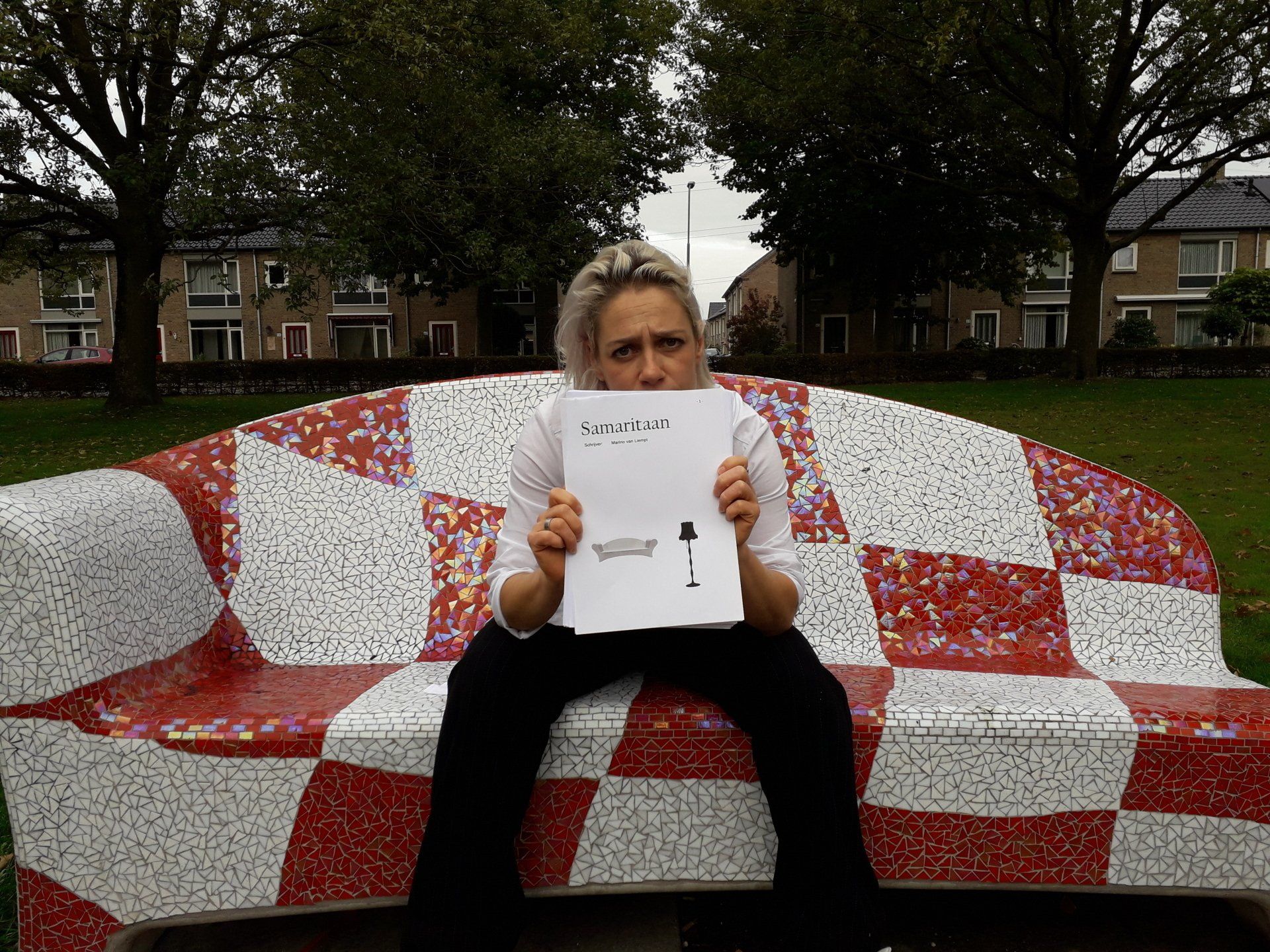 Een vrouw met een vel papier in haar hand zit bezorgd op een mozaïekbankje in een park.