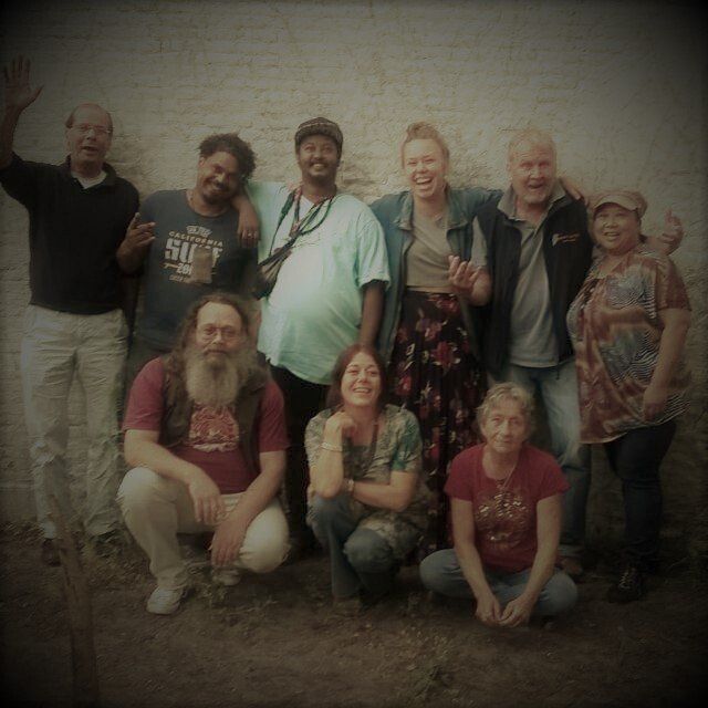 Een groep mensen poseert lachend in de buitenlucht. Sommigen knielen met hun armen om elkaar heen. Bewolkte, neutrale achtergrond.