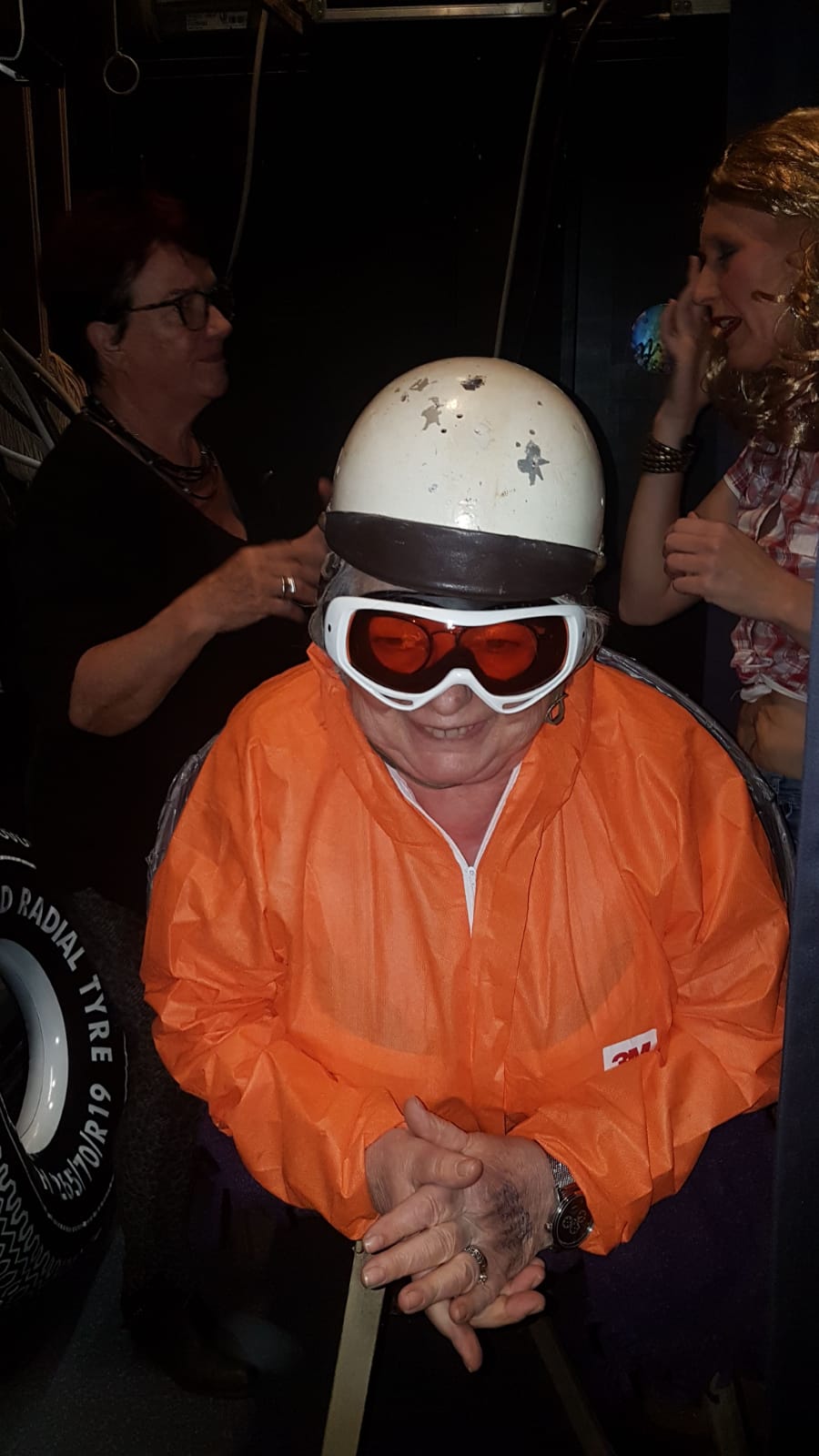 Een persoon in een oranje jas, helm en skibril lacht. Achter hem is iemand bezig met het in model brengen van zijn haar, op de achtergrond is nog iemand te zien.