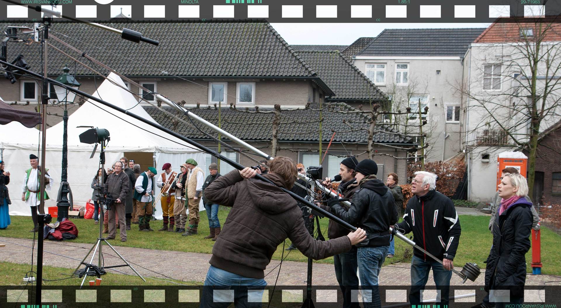 Filmploeg maakt buiten opnames: man met boommicrofoon, andere crewleden filmen, tenten en gebouwen op de achtergrond.