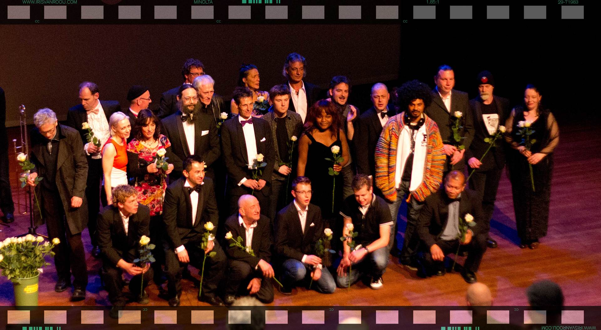 Een groep mensen staat op het podium, sommigen houden rozen vast en glimlachen. Formele kleding, theateromgeving.