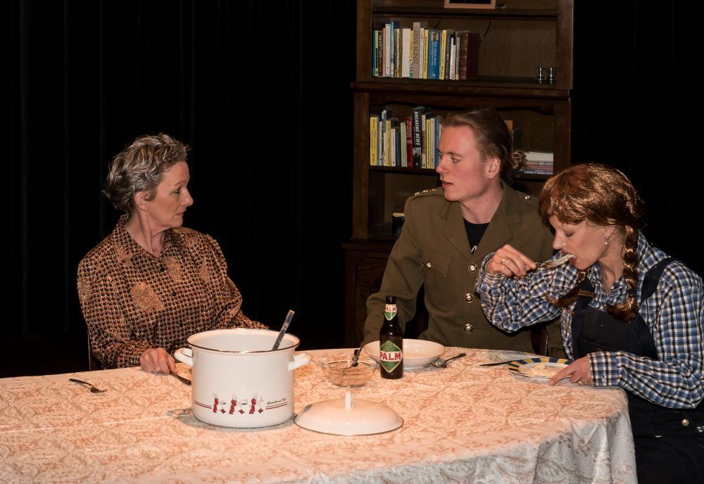 Drie mensen zitten aan een tafel, van wie één in uniform, te eten. Op tafel staat een pan, daarachter een boekenplank.
