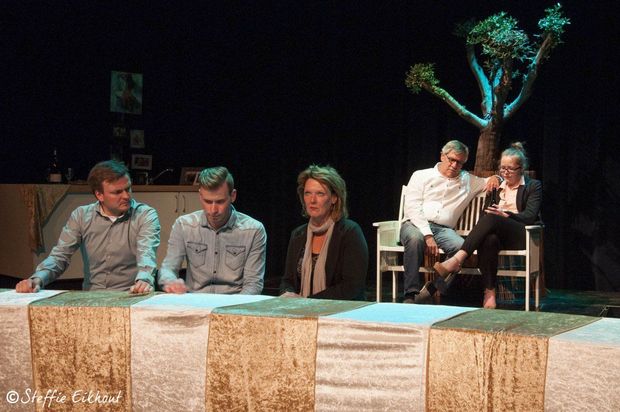 Vijf mensen aan een tafel op een podium; een man en een vrouw op een bankje onder een boom.
