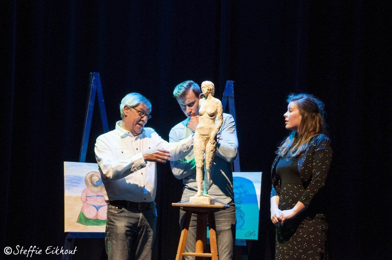 Drie mensen staan ​​op een podium en bekijken een naaktbeeld. Achter hen staan ​​twee schilderijen. Donkere achtergrond.