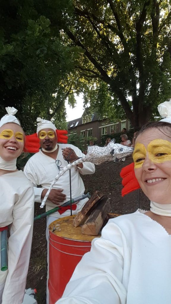Drie personen in witte kostuums met rode accenten en gele gezichtsverf staan ​​buiten bij een trommel.