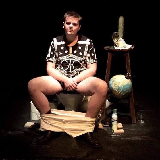 Een man zit op een toilet met zijn broek naar beneden. Een wereldbol, een kruk en andere voorwerpen liggen in de buurt.
