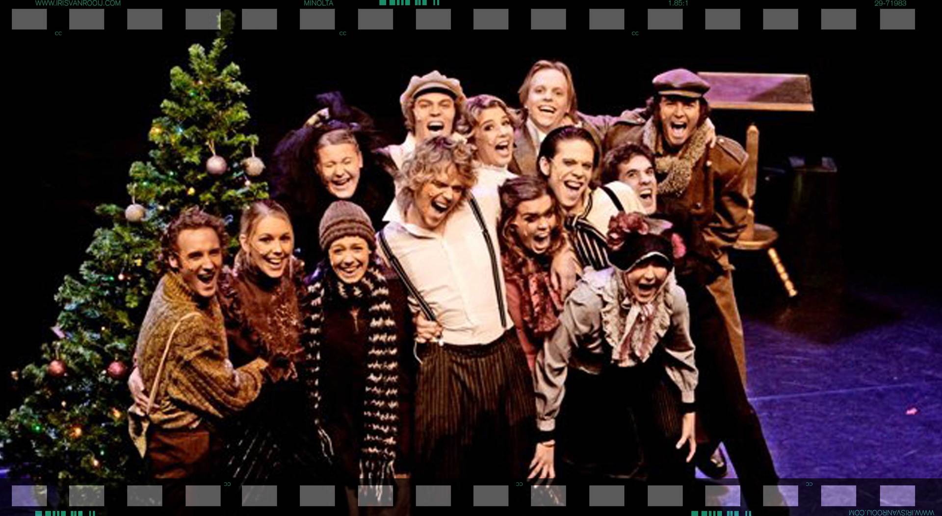 Toneelacteurs in historische kostuums en hoeden, lachend voor een kerstboom op het podium.