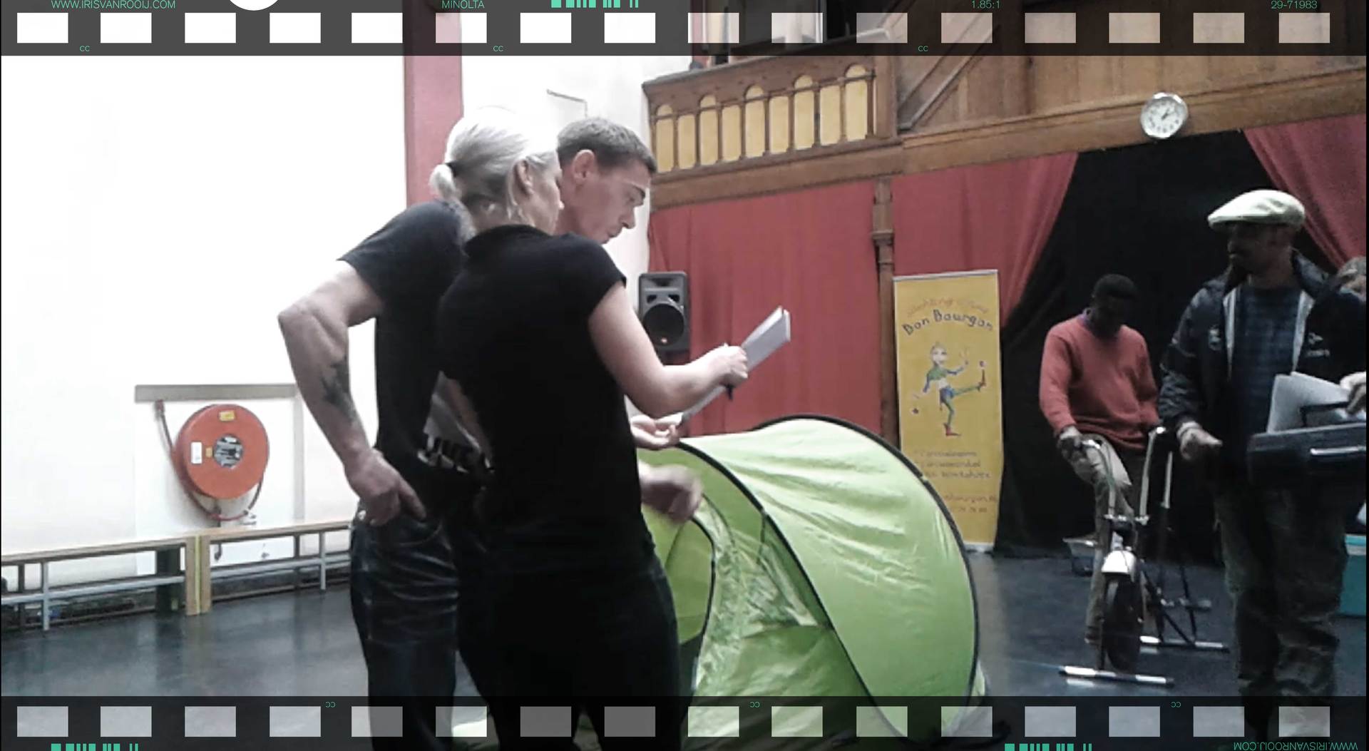 Mensen bekijken documenten naast een groene pop-up tent in een kamer, terwijl anderen op de achtergrond zitten.