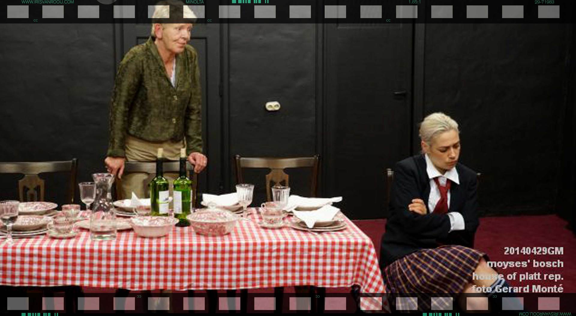 Twee vrouwen aan een tafel. De ene staat, de andere zit. De tafel is gedekt voor de maaltijd. Een rood geruit tafelkleed. Een donkere kamer.