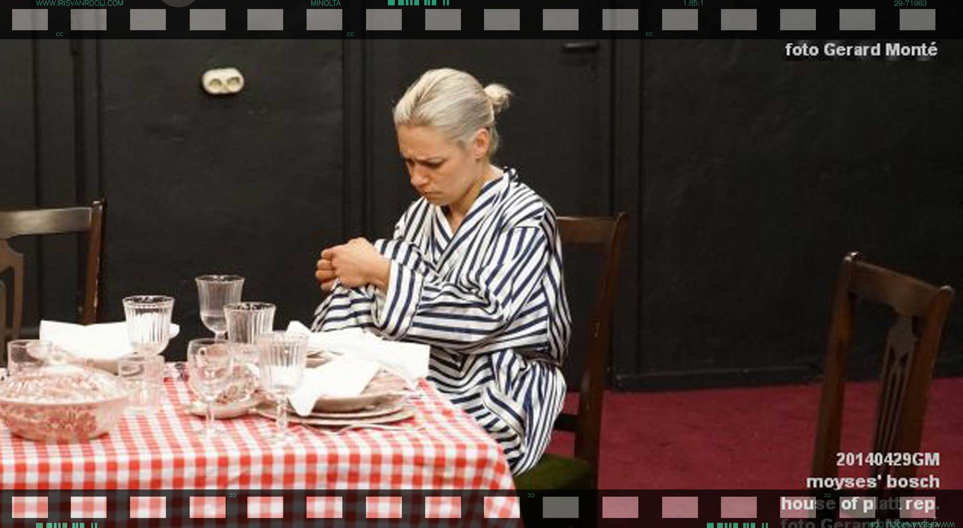 Een vrouw in een gestreepte ochtendjas zit aan een tafel met de handen ineengevouwen. De tafel is gedekt met servies en een geruit tafelkleed.