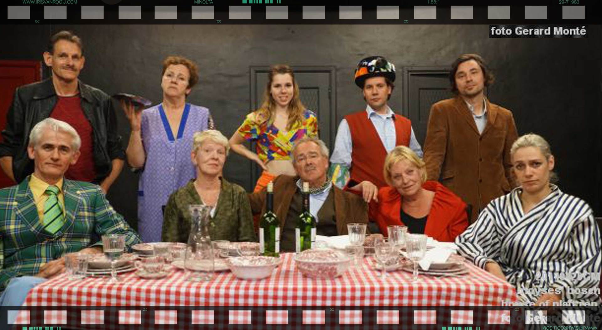 Een groep mensen staat rond een roodgeruite tafel te poseren. Ze dragen diverse kleding en sommigen kijken in de camera. De achtergrond is zwart.