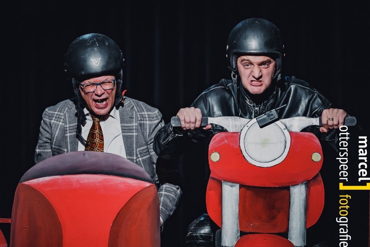 Twee mannen op een rode scooter, met helmen op. De ene in pak, de andere in een leren jas, trekken gekke gezichten. Donkere omgeving.