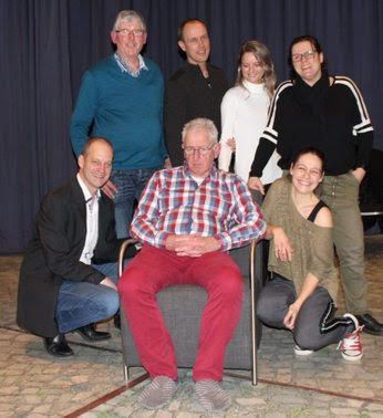 Een groep van zeven mensen poseert, een man in een rode broek zit, de anderen staan ​​of knielen. Donkere achtergrond.