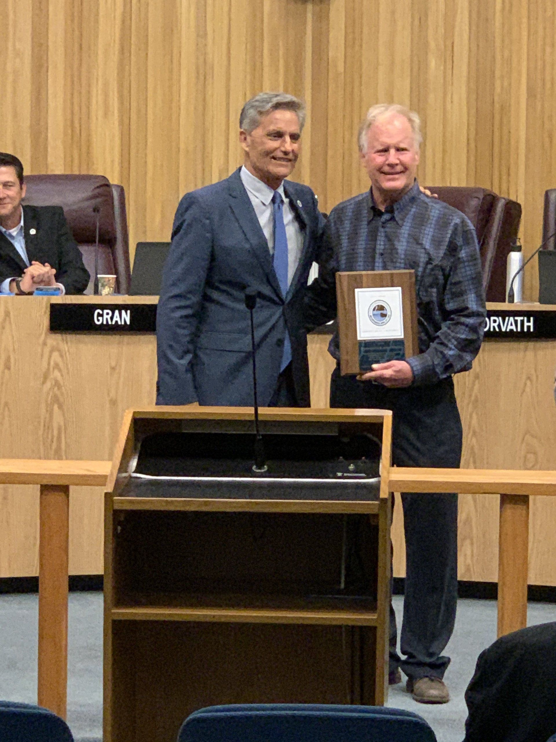 Two men posing in a council chamber, one holding a framed plaque, with officials seated behind them.