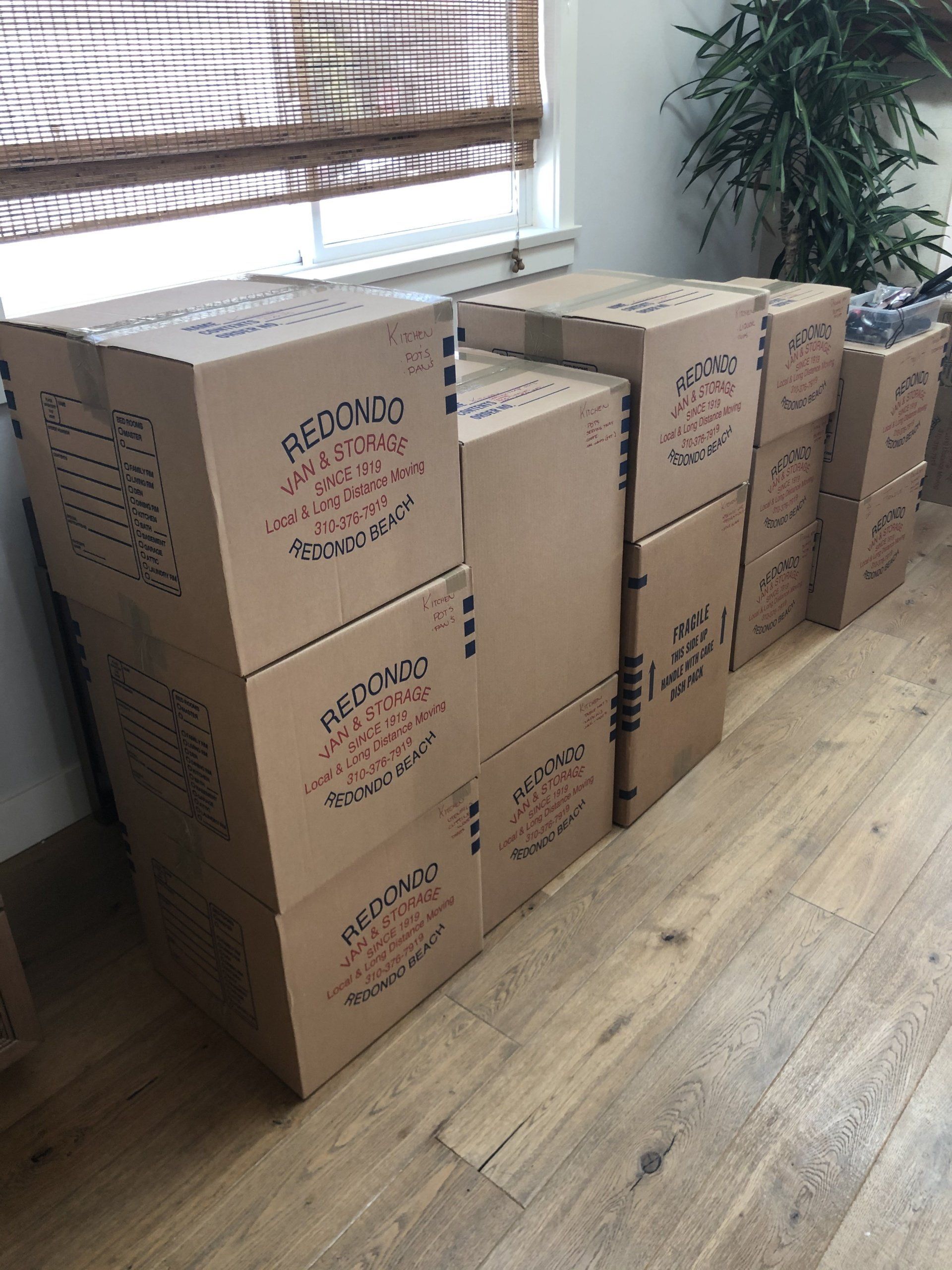 Stacked cardboard moving boxes in a sunlit room with wooden floors and a window blind.