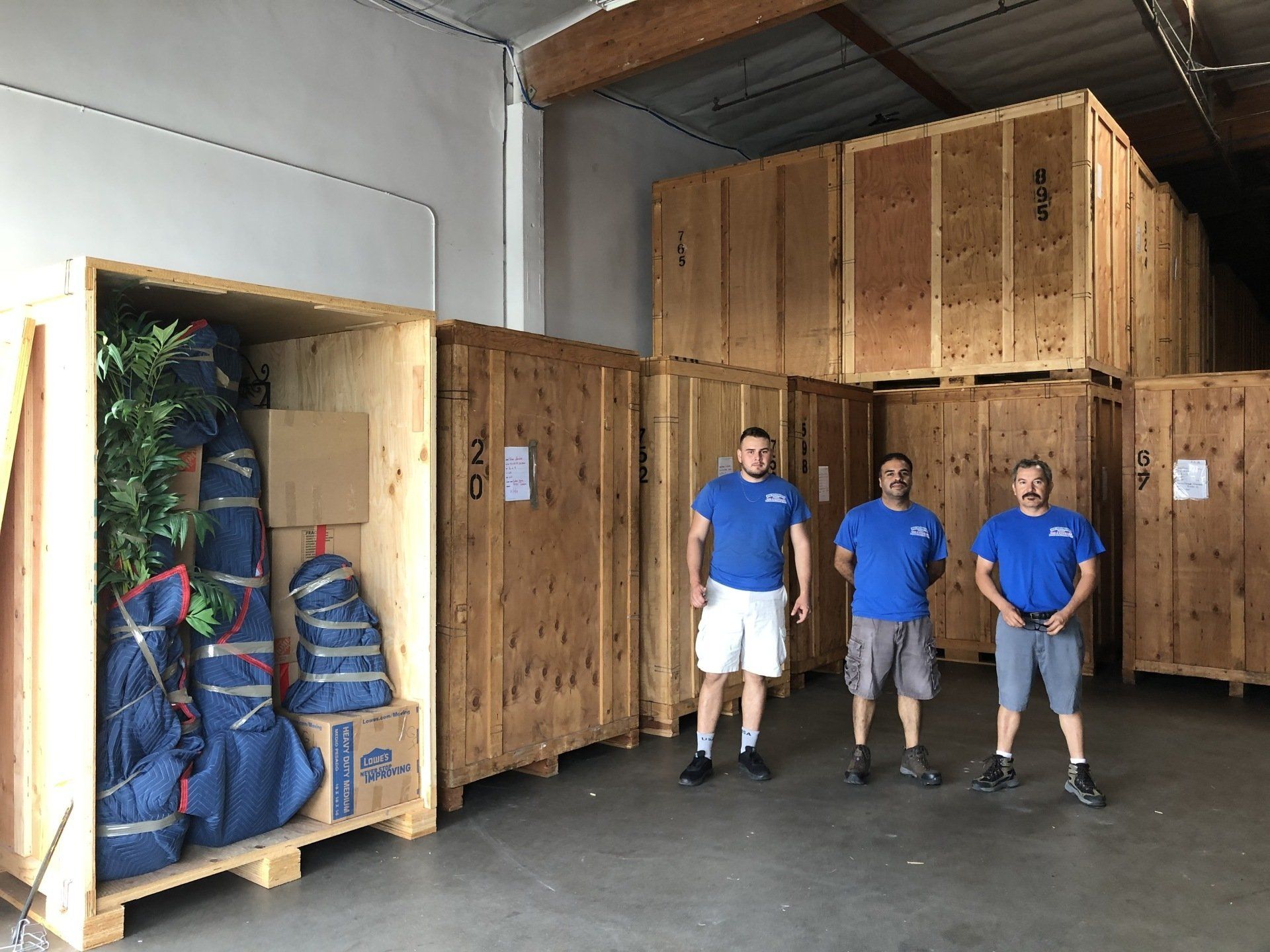 Three people in blue shirts stand beside large wooden shipping crates in a warehouse.