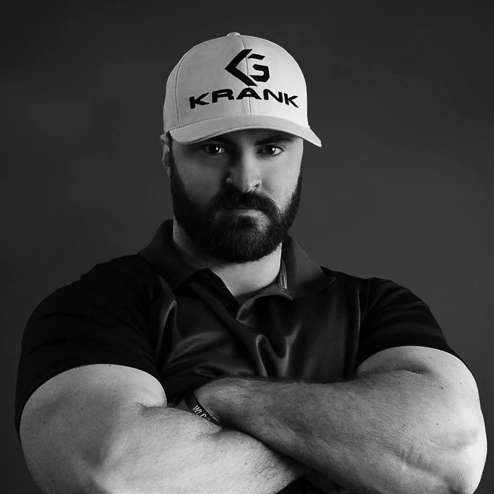 Man with beard and hat, arms crossed, looking at the viewer. Hat says 