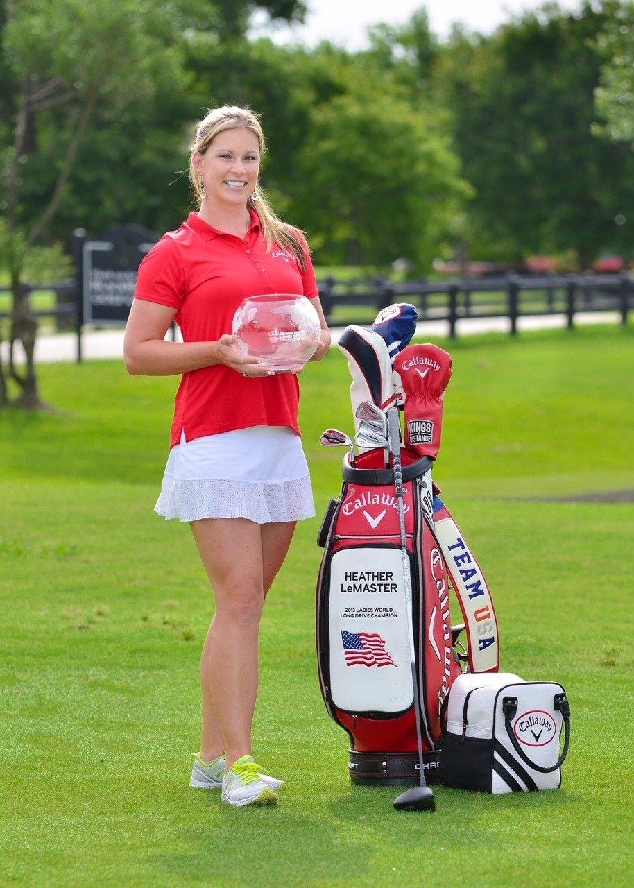 Golfer in red shirt and white skirt holds a bowl near her golf bag on a green course.