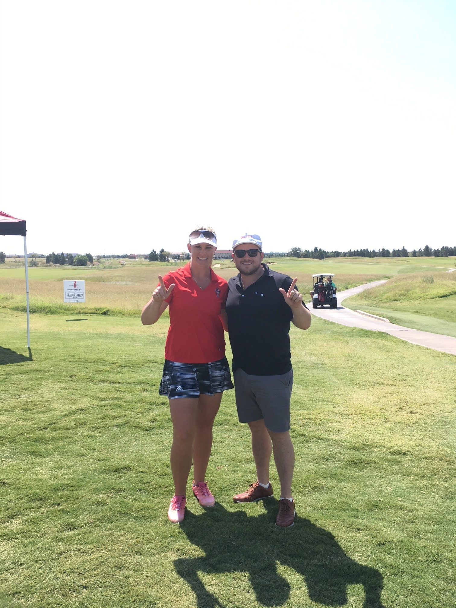 Two people on a golf course giving thumbs up. Woman in red shirt and patterned shorts, man in black shirt and shorts.