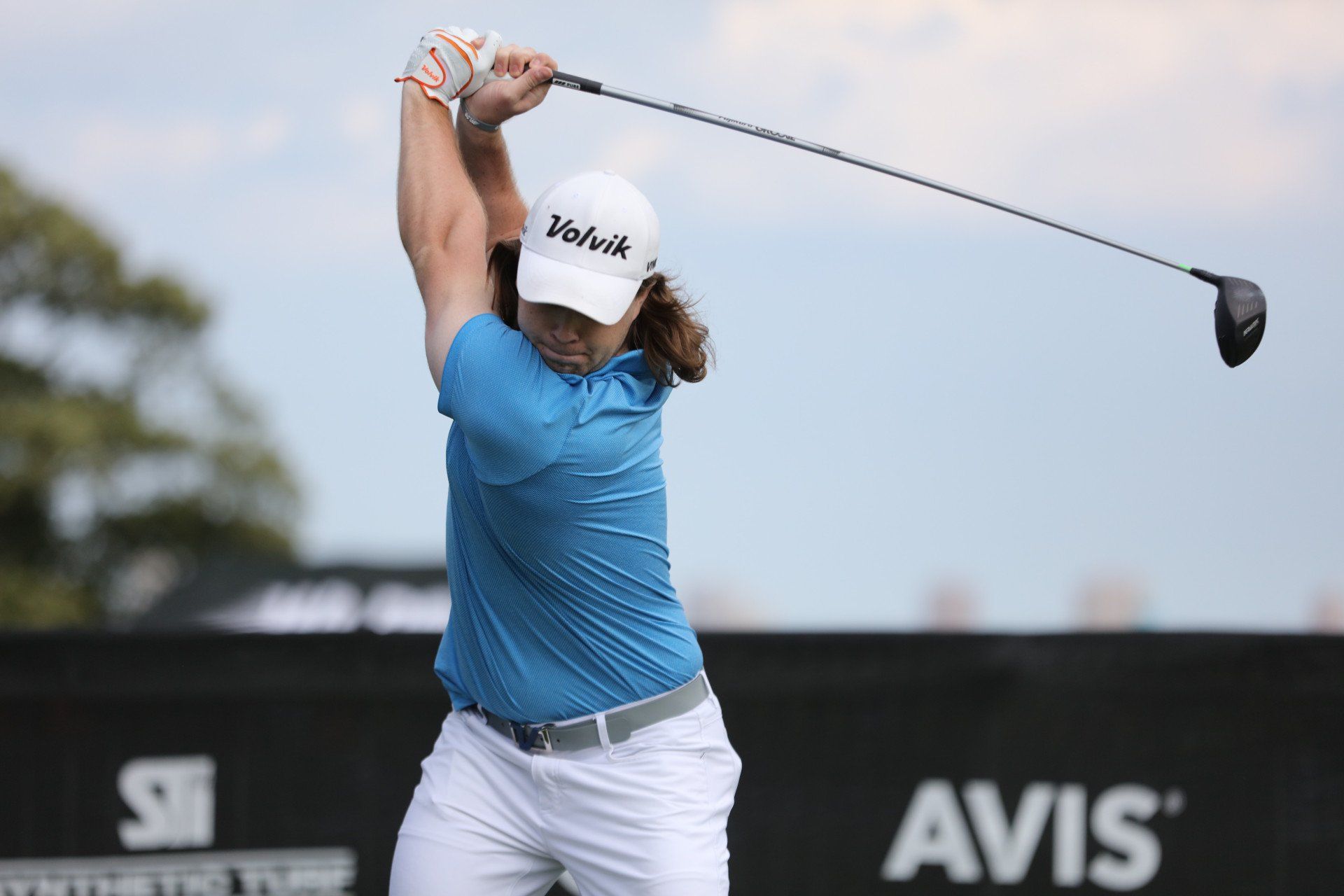 Golfer swinging club, blue shirt, white hat, driving on a course.