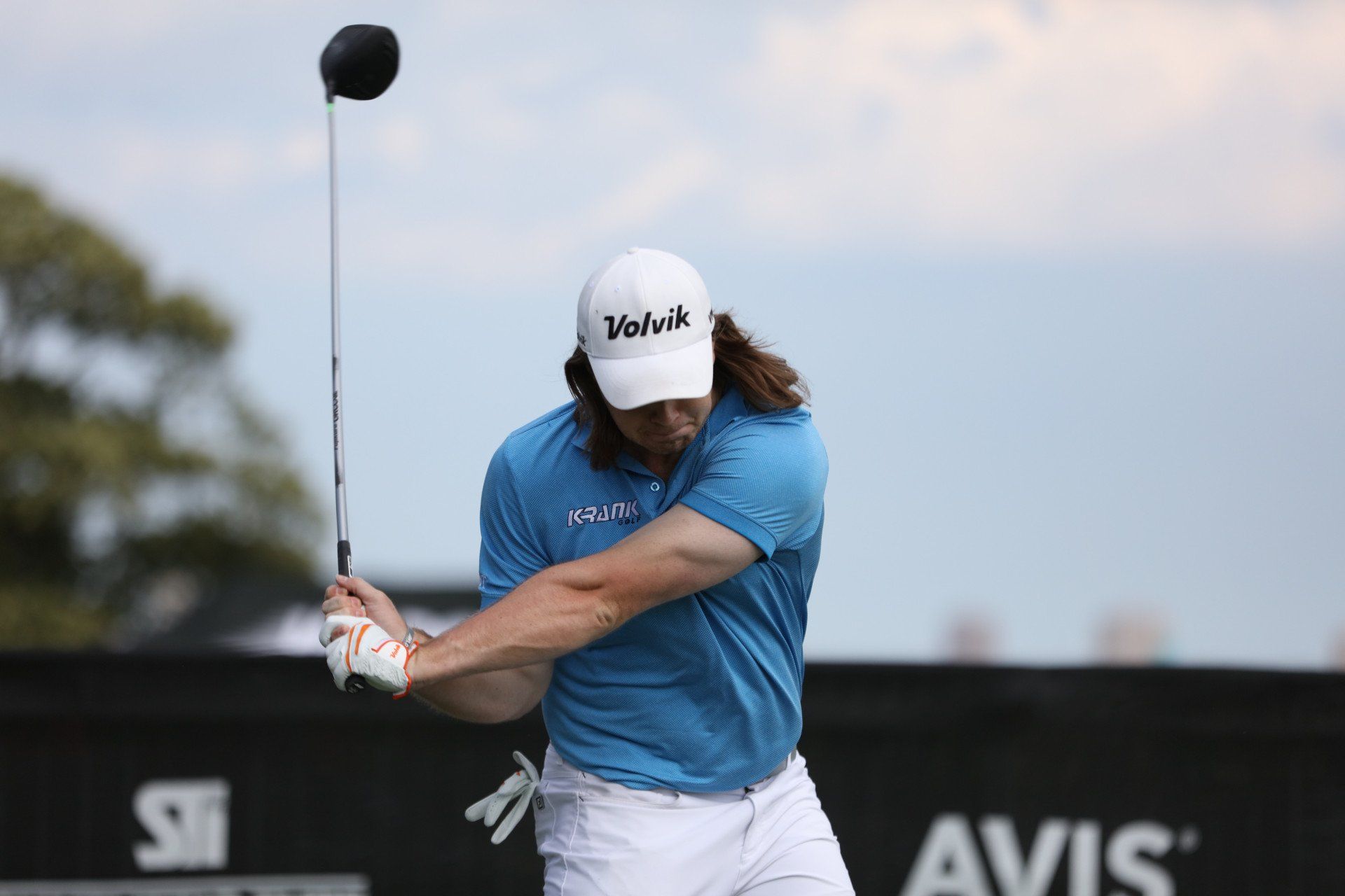 Golfer with long hair swings a driver on a course. He wears a blue shirt, white pants, and a white hat.