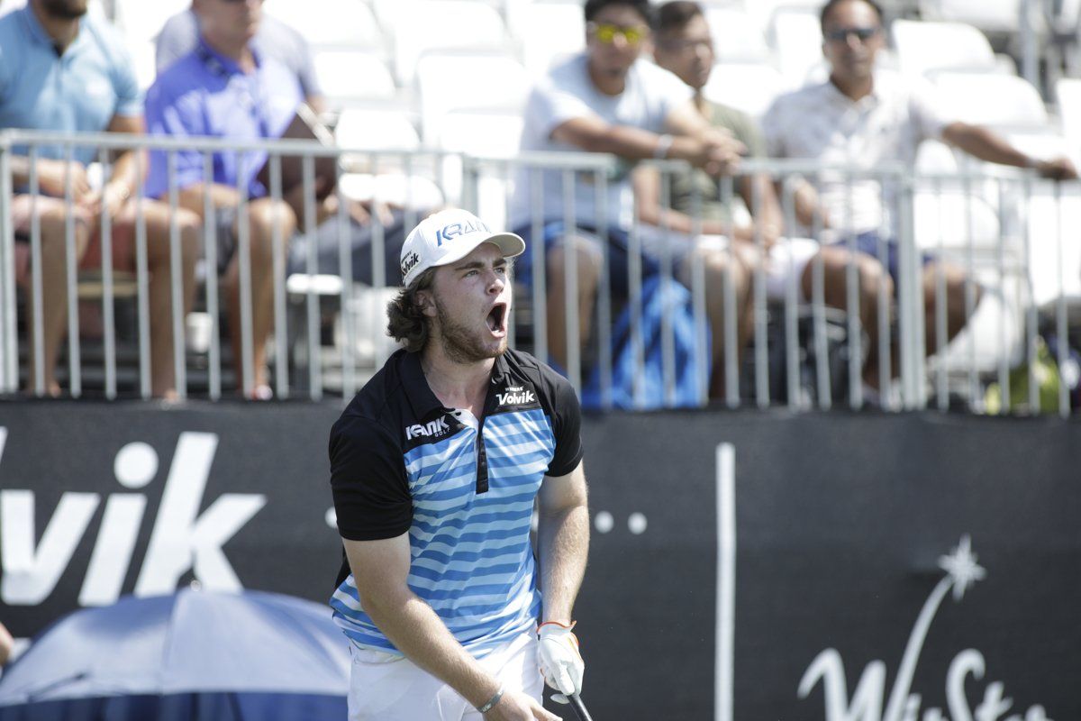 Golfer with open mouth, celebrating on course, wearing white cap and blue shirt, crowd in background.