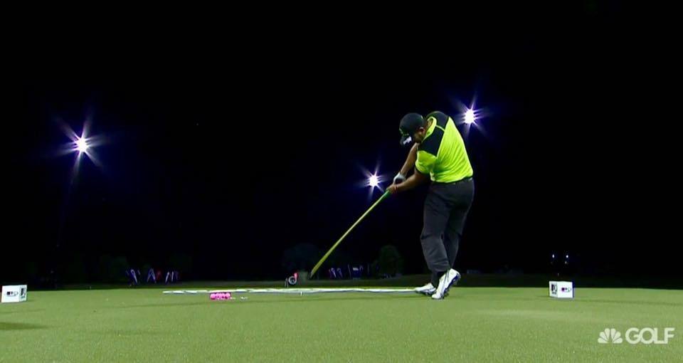 A man is swinging a golf club on a golf course at night.