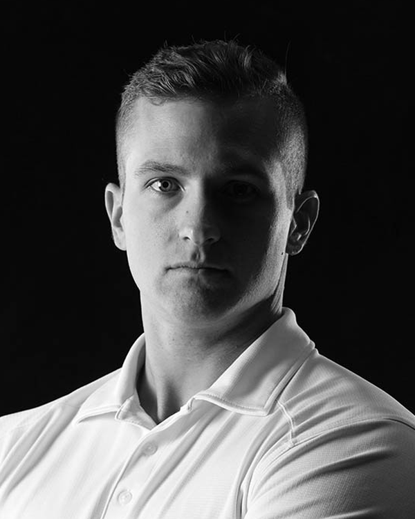 Man with short hair and white polo shirt in front of a dark background, looking directly at the viewer.