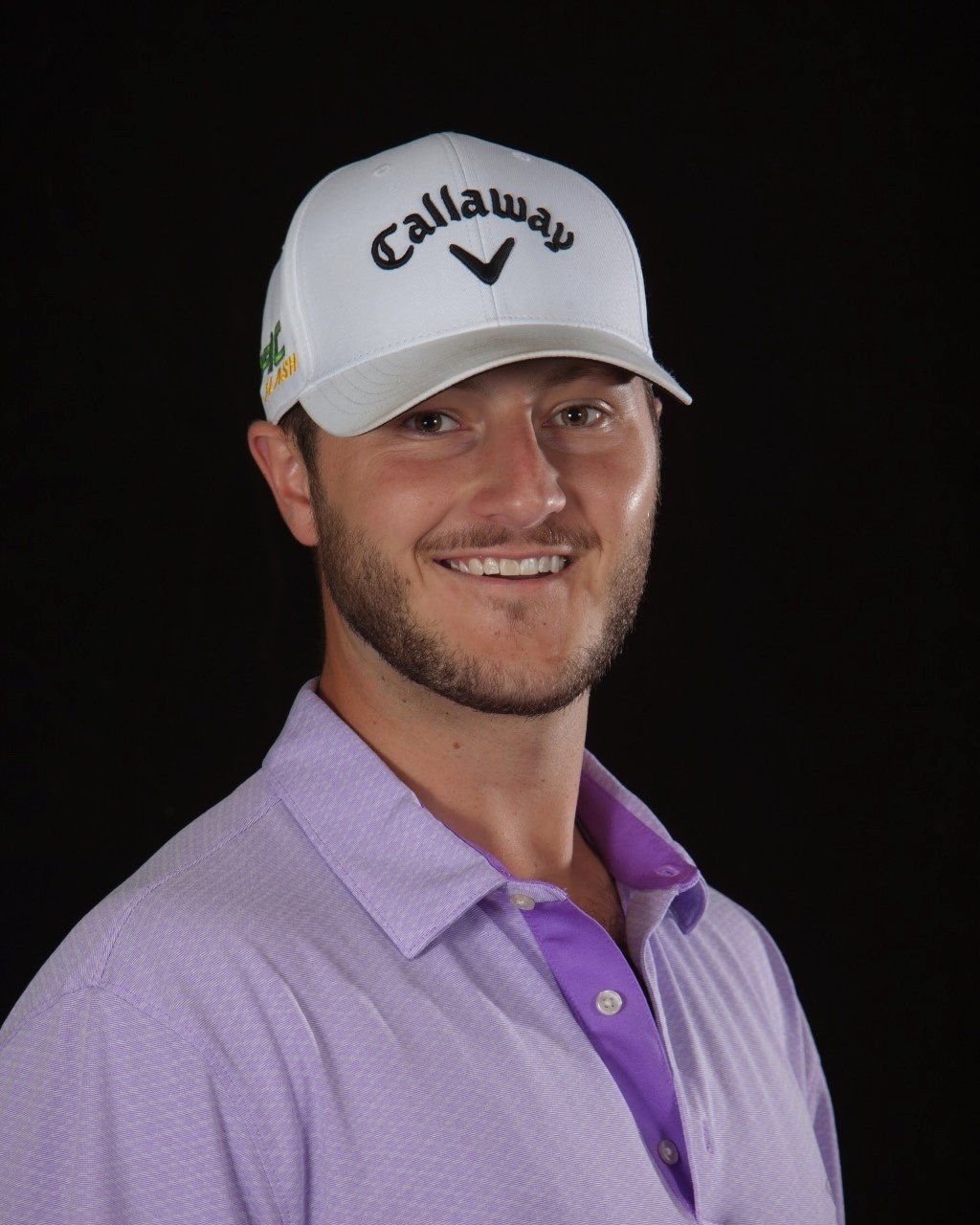 Man in Callaway hat smiles, wearing lavender golf shirt, dark background.