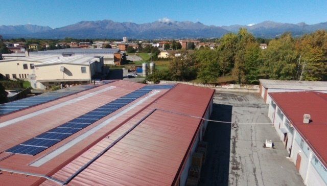 Veduta aerea di edifici industriali con tetti in metallo rosso, alcuni dotati di pannelli solari, sullo sfondo di montagne.