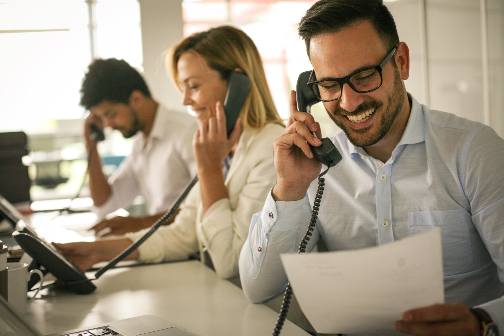 Three people at desks on phones; smiling man in glasses reading paper.