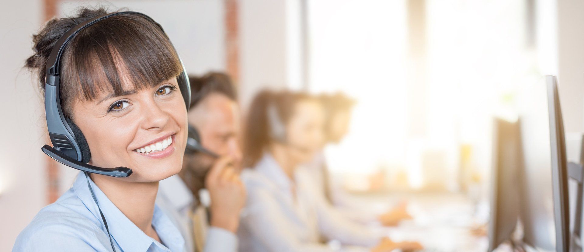 Woman with headset smiles at the camera in a call center setting.