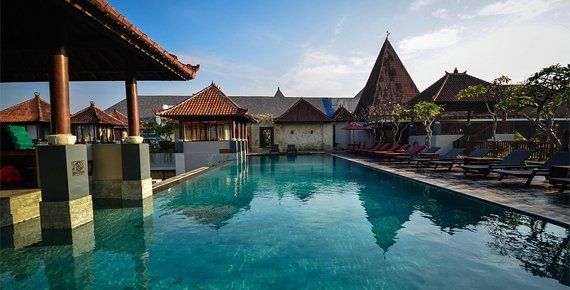 Pool with clear water reflecting sky, surrounded by Balinese architecture and lounge chairs.
