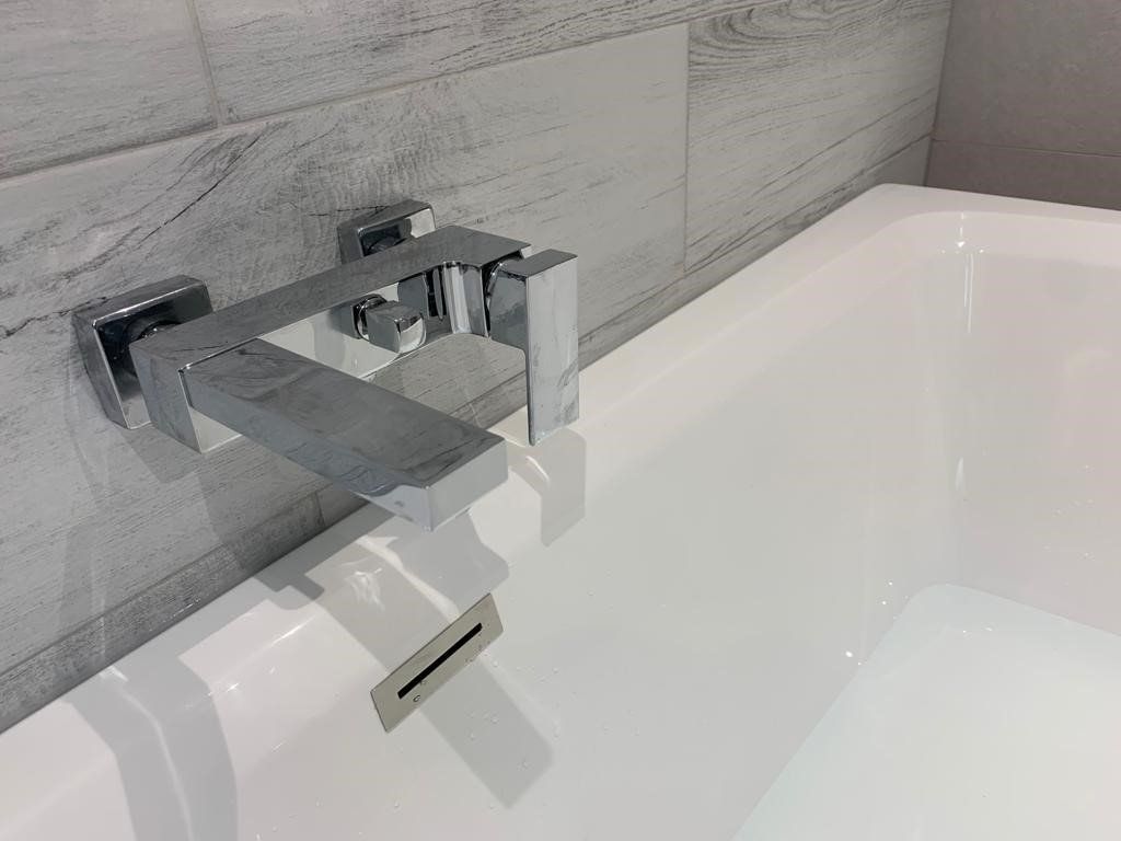 Modern chrome bathtub faucet and spout against gray tile, with white tub below