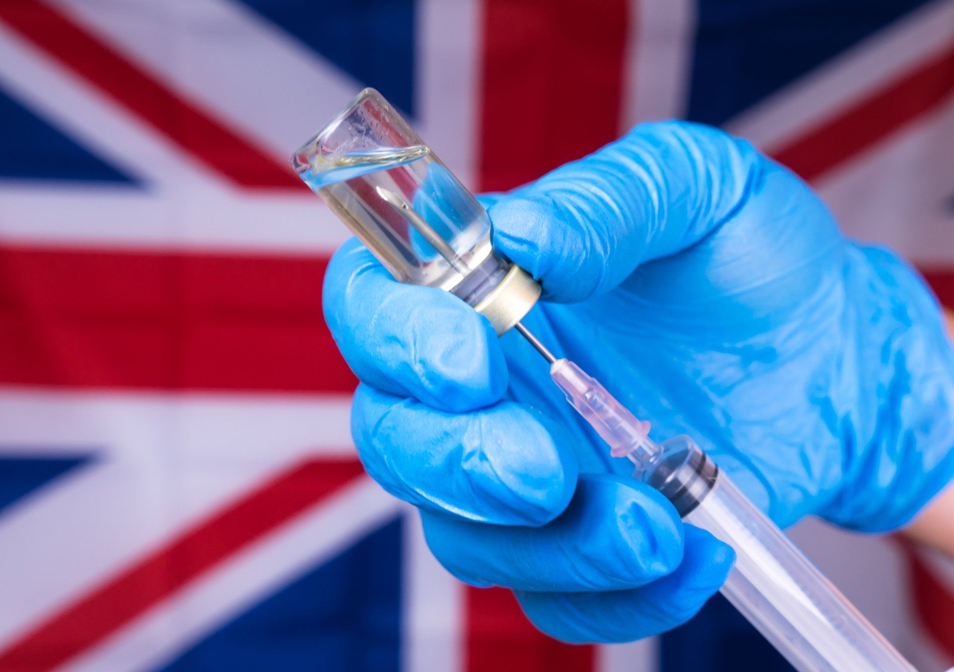Blue-gloved hand holding a syringe in front of a blurred UK flag