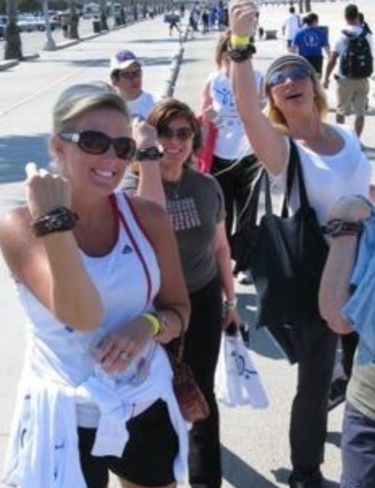 Group of cheerful people at an outdoor event, wearing sunglasses and raising their wrists to show off bracelets. They appear enthusiastic and engaged in what seems to be an awareness campaign gathering.